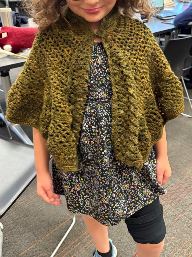 Front of the capelet, draped over a small floral print dress. Both front panels are visible, with the ruffle of the pineapple stitches falling over the model’s elbows. The fasteners sit perfectly at their neck, with the collar and triangles just falling short of their chin. The model’s smile is visible, and they’ve popped a knee forward for a sassy pose. 