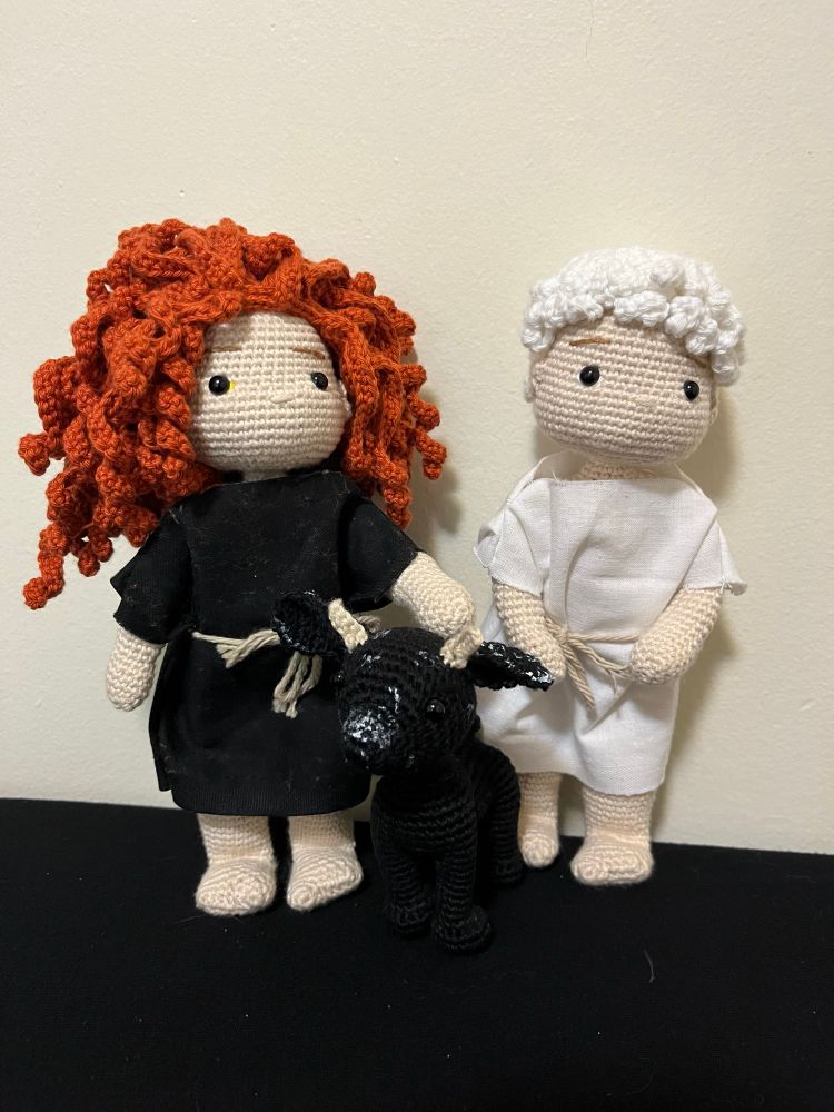 A trio of crochet dolls: one of Crowley, wearing a black tunic and with long curly hair, one of a little black crochet goat with white spots on her snout and ears, and one of Aziraphale wearing a white tunic