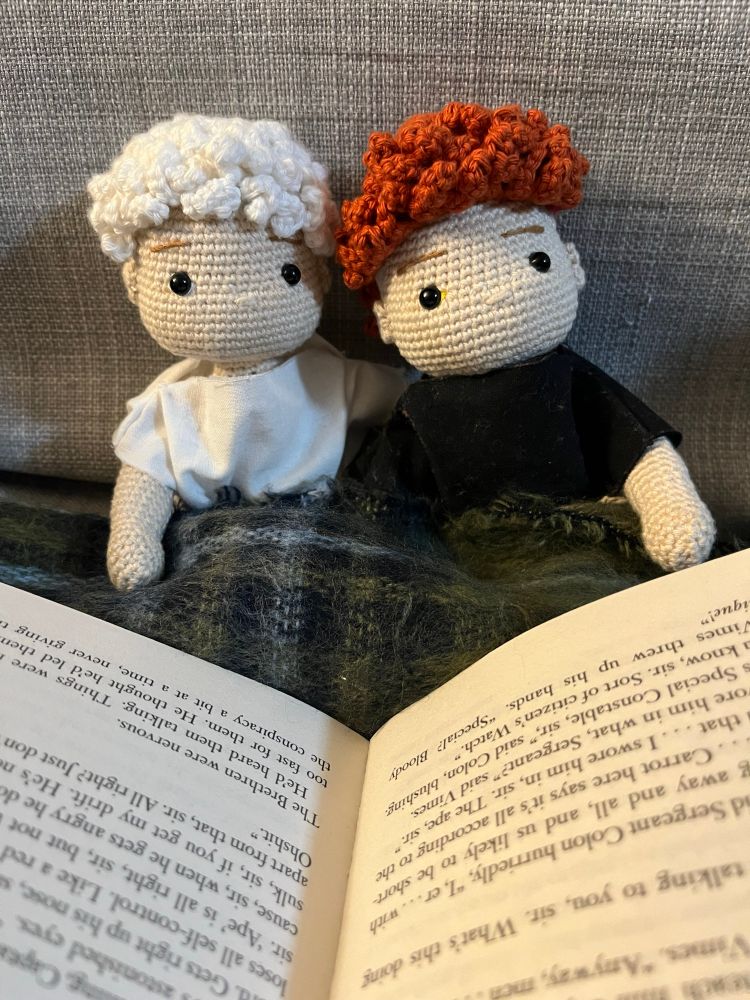 A pair of crochet dolls of Aziraphale and Crowley, sitting together with a tartan blanket over their laps and a human-sized book open in front of them