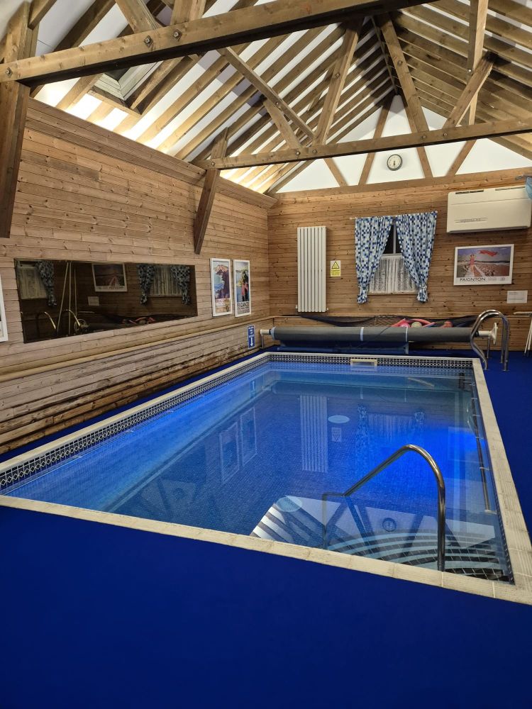 Picture of a very small pool housed inside a converted railway waiting room. The pool is blue and surrounded by blue flooring. The ceiling is vaulted with beams and the walls are clad in pine decorated with framed prints of vintage travel posters. This is the pool I went for a swim in. It was VERY warm and I had it all to myself, mostly, I did have to share it with @nibus.bsky.social but that doesn't count.