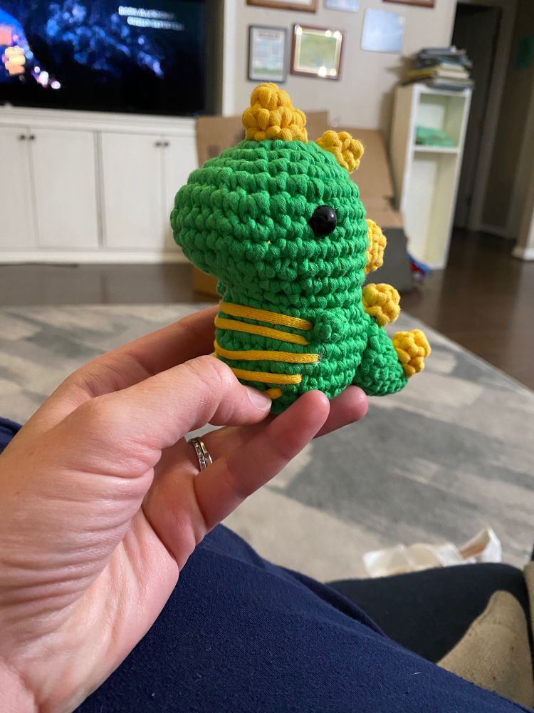 Stuffed crochet dinosaur (rawr!). Green body with 5 yellow spikes along its back and tail. Tension of the stitches were a little tight so its head pulls in one direction. Good Omens 2 is on the TV in the background. 