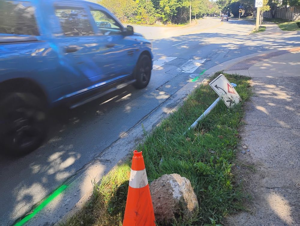 A crosswalk sign lies in the grass next to the four lane Lacewood Drive, near Braeside Lane. A blue truck speeds past. In the distance, a 50 km/h speed limit sign. Few drivers travel at this speed or slower