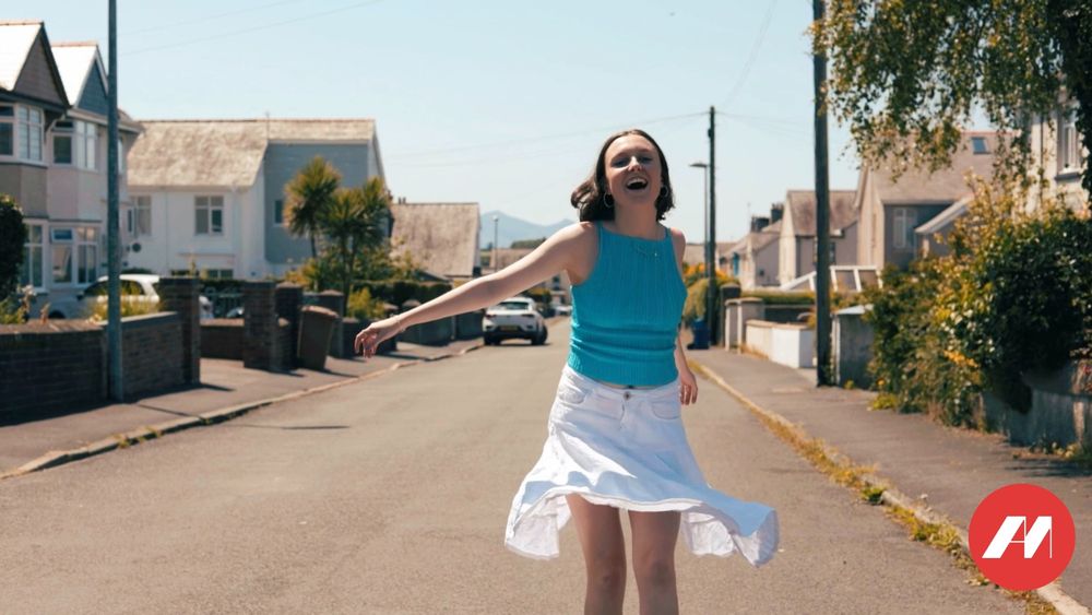 Still of 'Y Stryd' music video, featuring Alis Glyn in a blue top and white skirt, walking down a residential street.