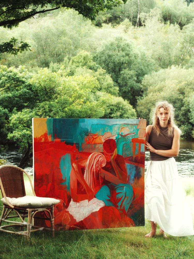 A person dressed in a white skirt and a brown top standing in front of a river. They are holding a large blue and red painting.