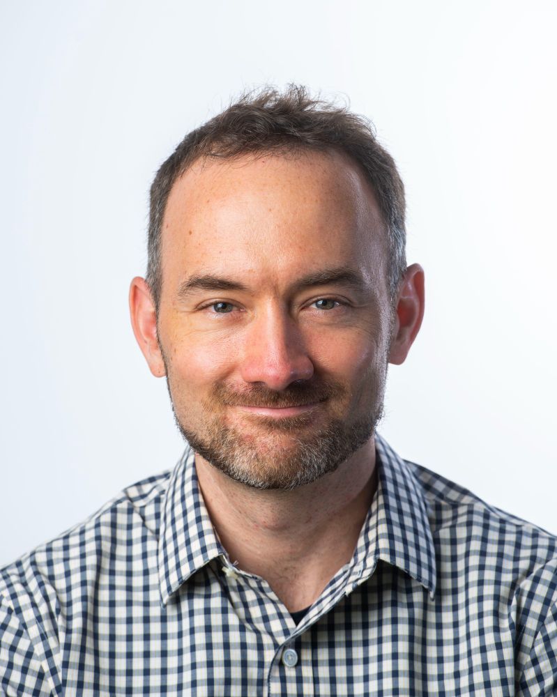 Portrait of a smiling person with short hair wearing a checkered shirt against a white background. The person in the image is Graham Neubig who will be presenting the lecture for this event.