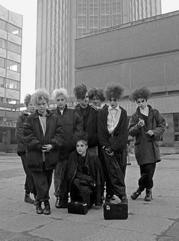 a group of goths, looking much like robert smith, in a modernist plaza