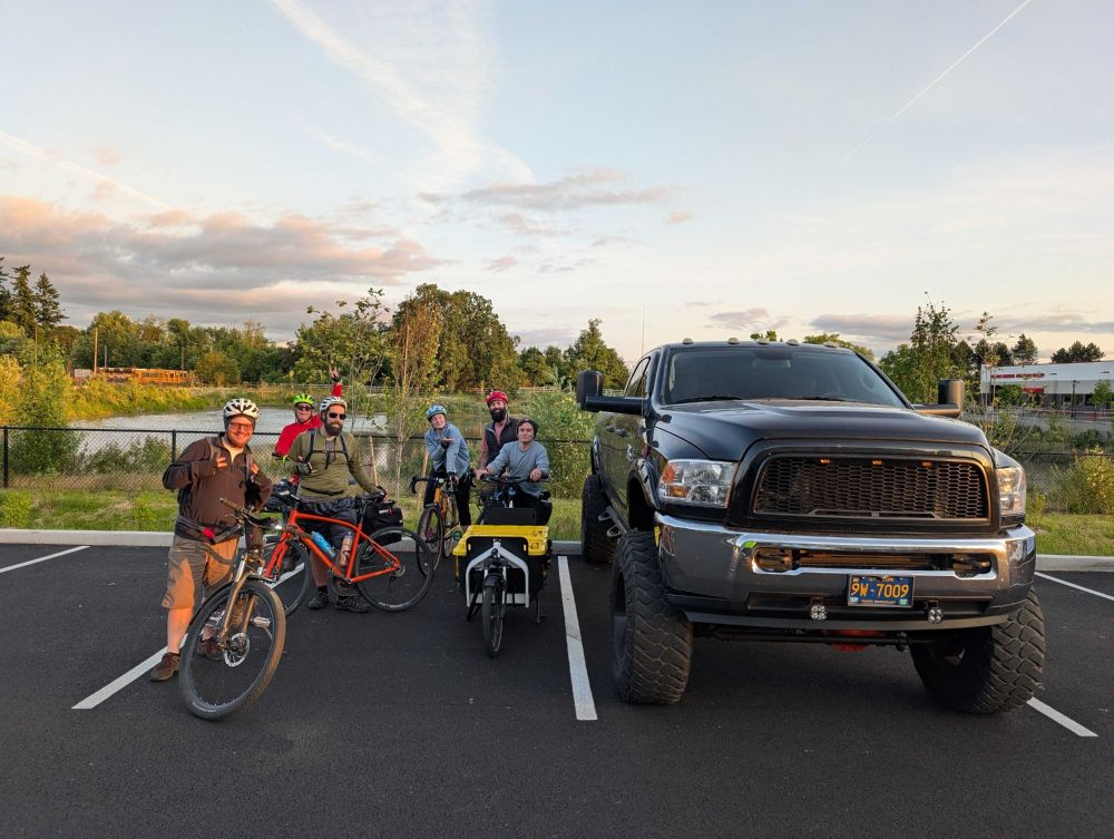 Six people and their bicycles looking small in a parking space next to one big pickup truck in a parking space.