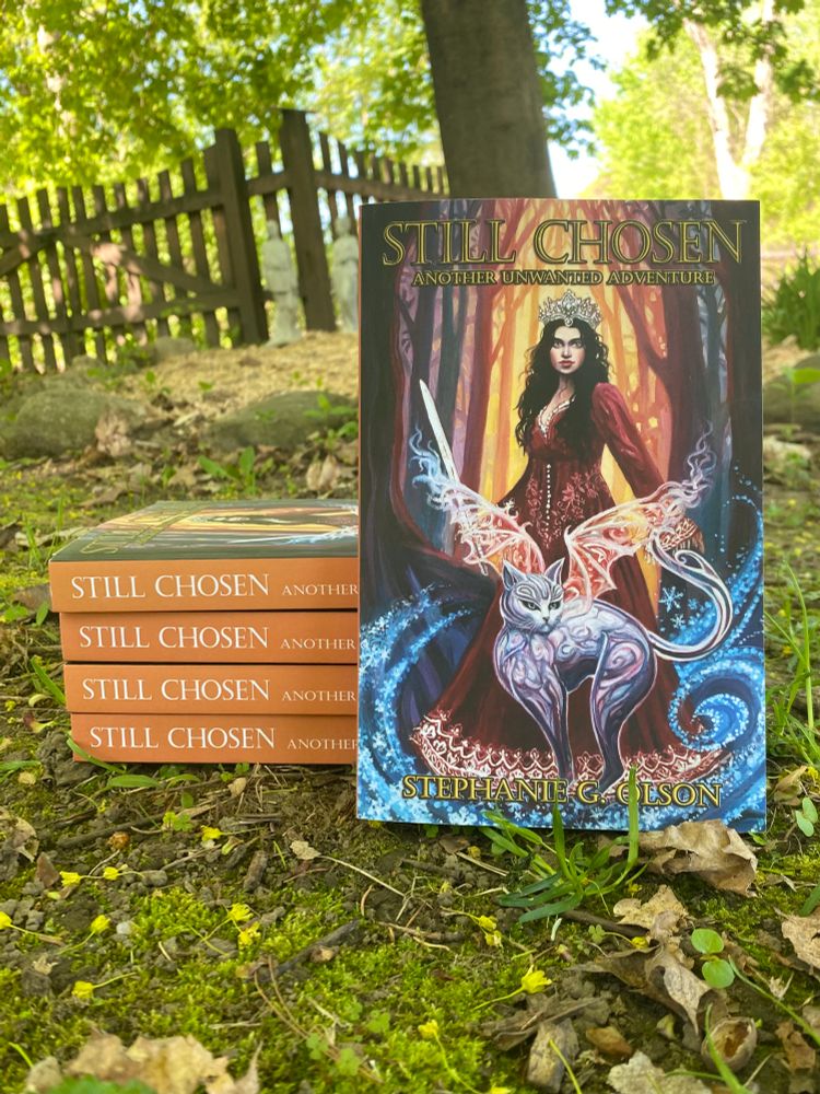 Stack of books outside under a tree, with one propped up to show the cover. Cover reads "Still Chosen: Another Unwanted Adventure" and features a woman in a dress and crown holding a sword, and a large silver cat with wings.