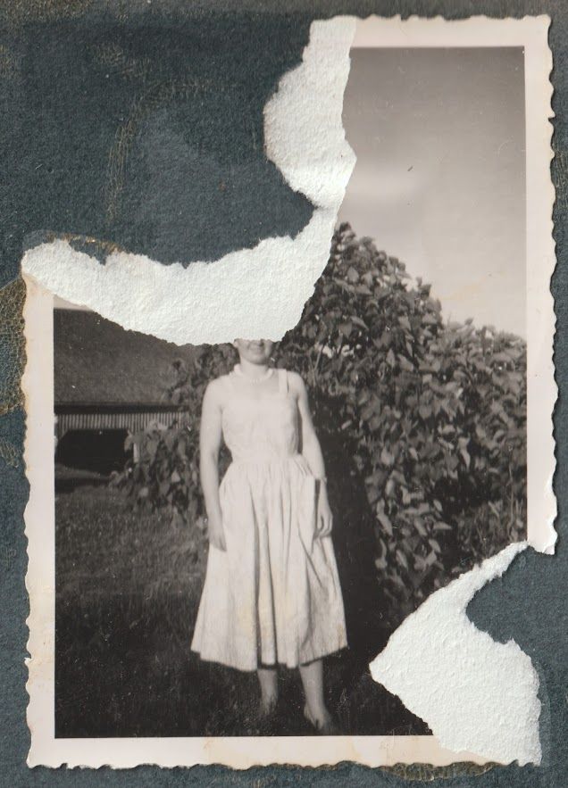 Vanha valokuva, mustavalkoinen, repeytynyt, kuvassa on nainen leningissä, kesä, hän on sireenipuska edessä, poseeraa, taustalla näkyy piharakennus