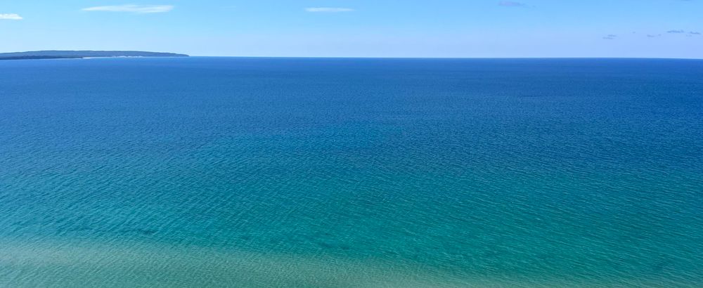 Point Betsie and Platte Bay, from Empire Bluffs; Empire, MI

Color image, unbroken and calm blue/blue-green waters of the larger Lake Michigan. At the horizon left is a distant strip of land. Sky is hazy blue with white cloudiness beyond the horizon.