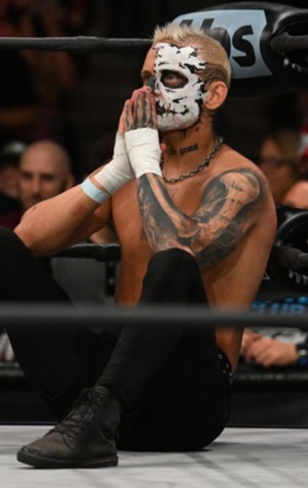 Blonde man with a half painted face seated against ring turnbuckles with hands clasped in front of his face 