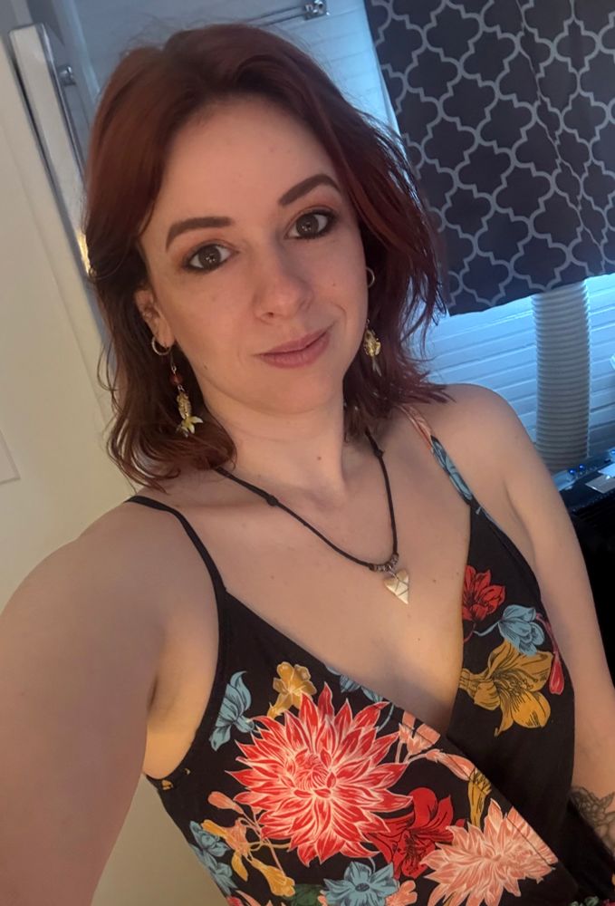 A portrait selfie of me, showing my head and shoulders down to my waist. I am wearing a black dress with spaghetti straps and a V neck. It has an abstract floral pattern consisting of pink, blue, peach, and mustard yellow flowers which scroll along in an organic flowing pattern. I am wearing a shark tooth on a black cord with brown beads around my neck. My hair is red and shoulder length. I am also wearing gold koi fish dangle earrings. 