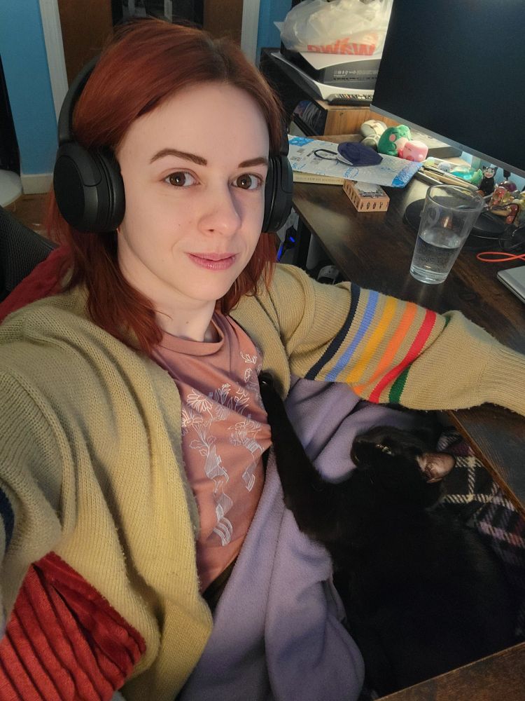 A selfie of me sitting at my computer desk with my cat. I have red hair and am wearing a pink and white shirt and tan open front sweater with rainbow stripes on the sleeves. My red electric heated blanket is around my shoulders. On my lap is a light purple and dark purple plaid blanket, and my Kat Killian is sitting on it, with her left arm outstretched and paw on my chest.