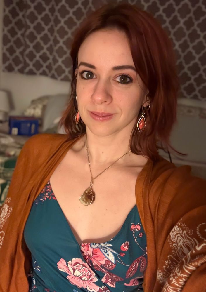 A selfie of me, curiouscat. It is a portrait shot of head to chest. I am wearing a teal dress with a pink and white floral pattern and over top of that an orange shawl with white bohemian details. I am wearing an orange citrine rock on a gold chain around my neck because I like rocks! I have diamond shaped dangle earrings which are gold on the outer rim and salmon pink in the middle. My hair is shoulder length and is copper red.