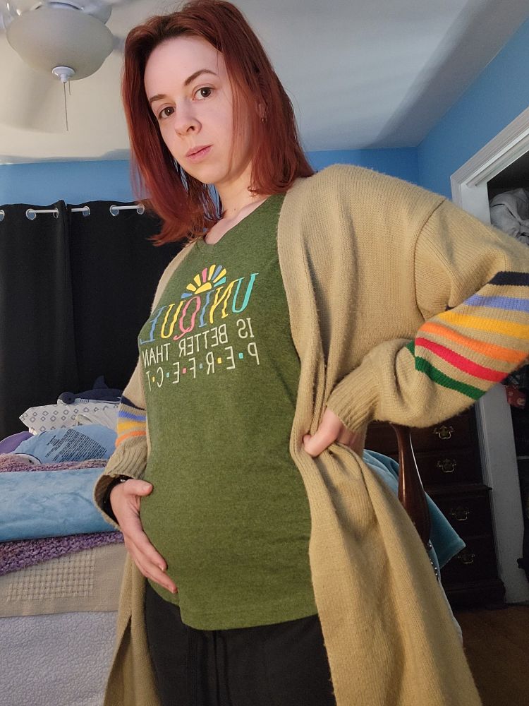 A photo of me done in the style of a pregnancy announcement photo with my right hand on my belly which is much bigger than normal, but actually, it's just bloating from my sterilization surgery. I do, however, look pregnant. I am in the foreground wearing a medium green shirt and black sweatpants with a long tan sweater over it which has rainbow stripes on the sleeves. My bed is in the background which I crashed into immediately after this. 