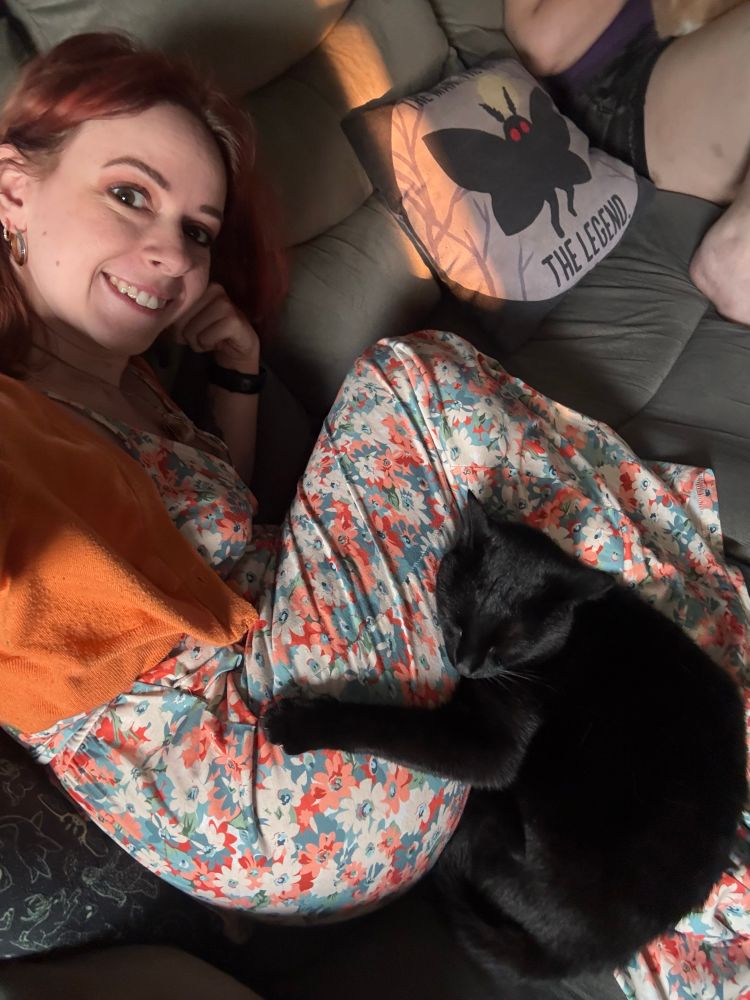 A picture of me wearing a floral print dress curled up, with Kiki the black cat curled up between my legs and butt. One of her arms is extended and resting on my hip like she’s hugging me