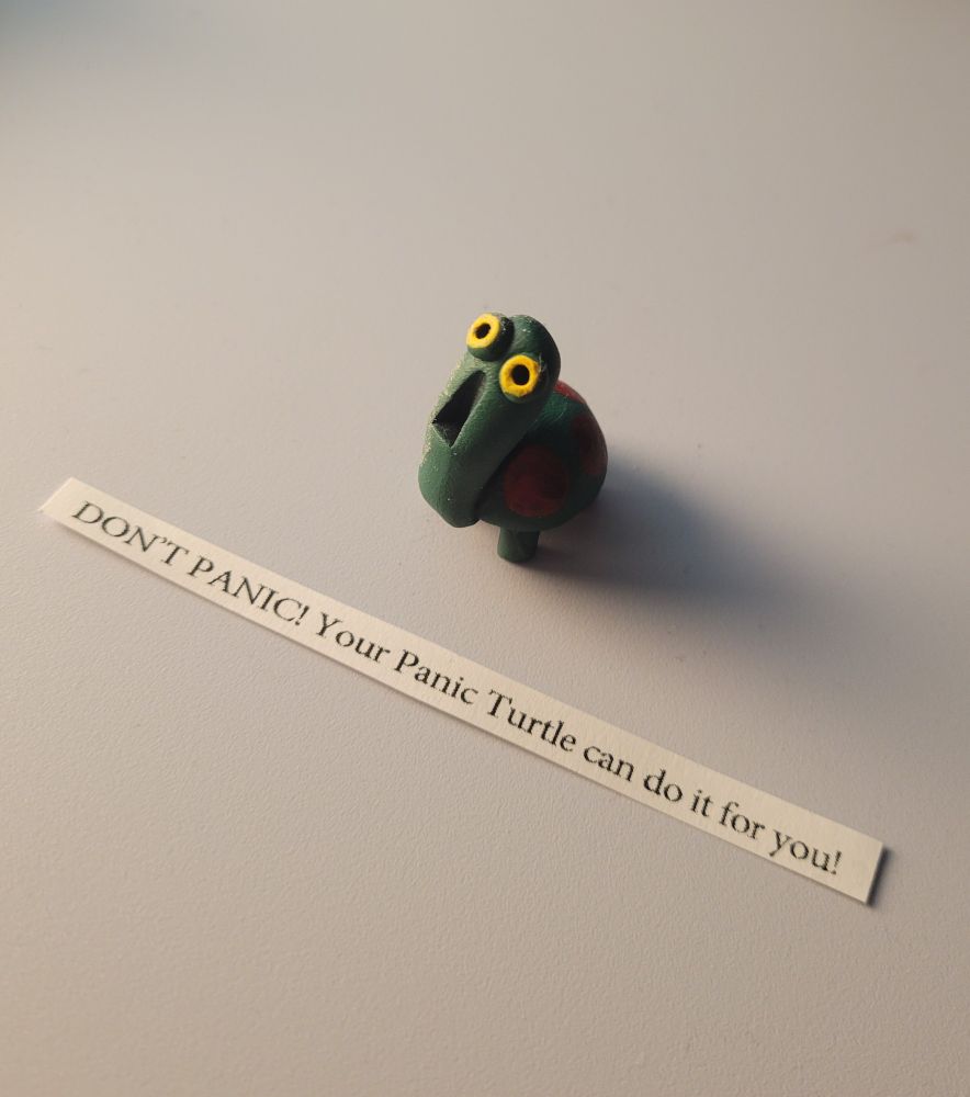 A small clay statue of a stylized cartoon-like green turtle with red circular spots on the shell. His yellow eyes are bugged out in shock, and his mouth is agape. There is a slip of paper, which came with the turtle that reads "DON'T PANIC! Your panic turtle can do it for you!" The background is plain white, because I balanced him on top of my playstation to take the photo, which is white.