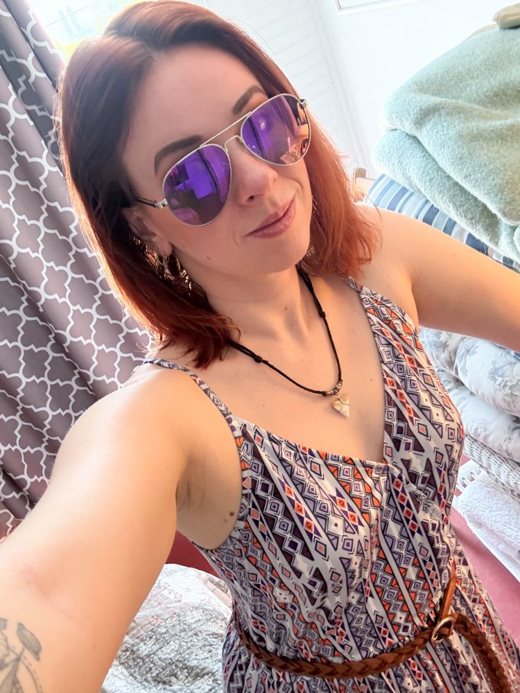 A selfie of me. I’m wearing a dress that has spaghetti straps and a V neck with a geometric pattern of mostly diamond shapes in purple, orange, and white with a brown braided belt at the waist. I am wearing matching purple aviator sunglasses and a shark tooth on a black cord with brown beads around my neck. My hair is shoulder length and ginger red. 