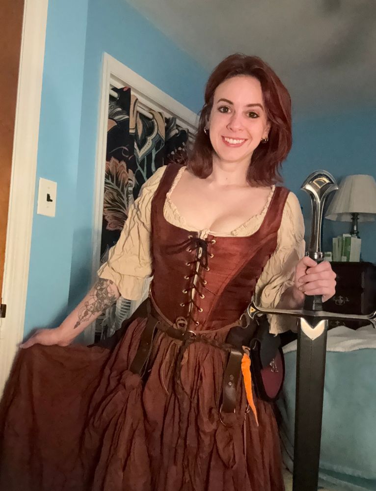 A photo of me from about my head to my knees in one of my renaissance fair outfits that I’m building this season. I have a light tan shirt with a deep copper colored bodice that laces in the front and a long rust brown skirt. I am wearing a braided thin brown leather belt with skirt hitches, an orange worm on a string I received last year and now hang from my belt and a red and black leather bag which attaches to the belt. My hair is red, shoulder length, and layered. I have a tattoo visible on my right forearm (our left looking at it) which is an abstract compass with a Paragon wing from Mass Effect and planet and floral motifs. In my left hand (our right viewing it) I am holding my replica of Anduril, Aragorn’s sword from Lord of the Rings, which was gifted to me by my partner GawkingGeek, and of which I am very proud. The sword is so long it comes up to my chest when I rest the tip on the ground but I’m holding it up a little so it’s shoulder height. I am standing in my bedroom. The walls are sky blue and in the background I have black curtains with large abstract many colored floral patterns and my big wooden dresser.