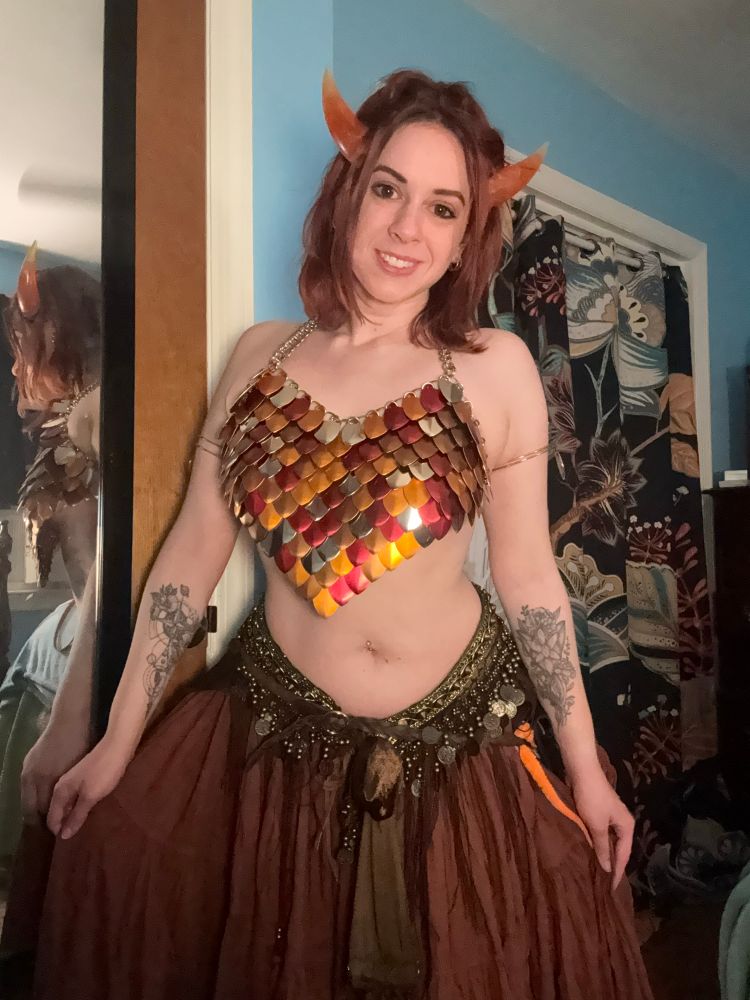 A photo of me in an outfit I’m gearing up for this season’s ren fair, from my head to a bit above my knees. I wore it last year but I’m always adding to my builds. I am wearing a halter neck dragon scale maille top which has red, orange, gold, and silver scales. I am wearing a rust skirt low on my hips with a fancy belly dancing scarf with little coins that jingle when I move. I am wearing gold bands around my upper arms. I have shoulder length layered copper red hair and am wearing a pair of pointed orange horns on my head mixed into my hair to be a bit more natural. For fair day I will probably try to do some more styling. I am holding the skirt out a bit with my arms down and slightly outstretched and you can see both forearm tattoos, space themed abstract compass with a paragon wing from Mass Effect on the right (our left) and an Evenstar from the Lord of the Rings movies on my left (our right). I’m standing in my bedroom. The walls are sky blue and I have black curtains with many colored abstract floral patterns.