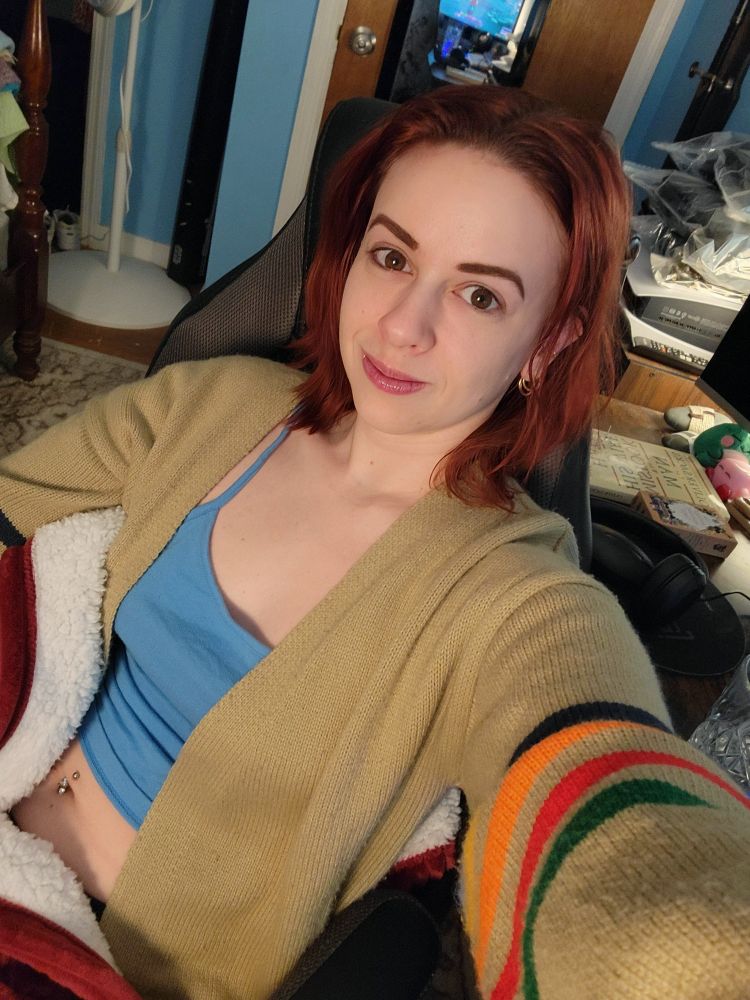 Selfie of me, curious cat, sitting at my computer desk in an office chair. I have red hair and am wearing a cropped blue camisole with a tan sweater that has stripes of color on the sleeves. Around my waist I have a red and white electric blanket.
