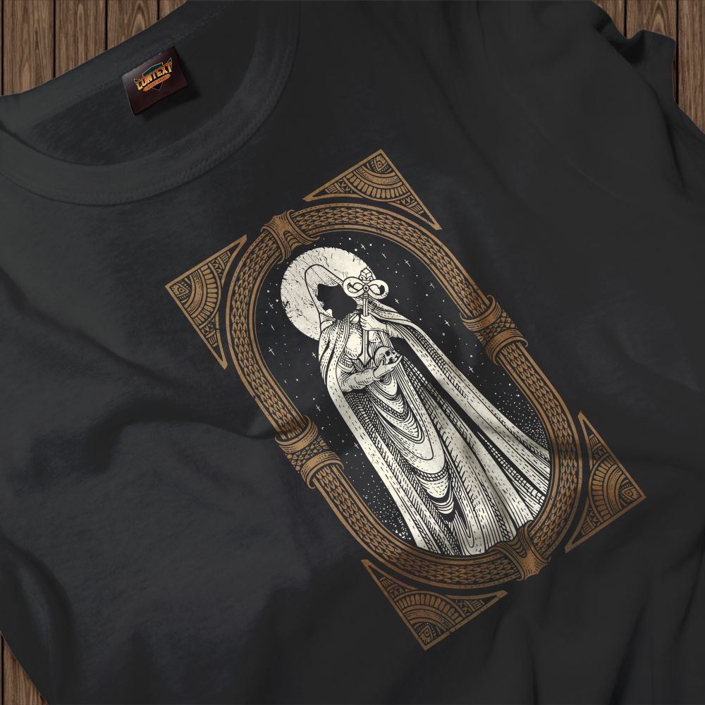 A dark gray T-shirt is laid flat, centered with a stunning monochrome illustration of a robed figure that evokes a witch or priestess. The character is highly detailed, holding a bundled object, and stands against a background of stars and a crescent-moon halo. The entire scene is framed by an intricate, vertically oriented rectangular border featuring repeating geometric and tribal-like patterns in bronze and brown, giving the design an antique, mystic aesthetic.