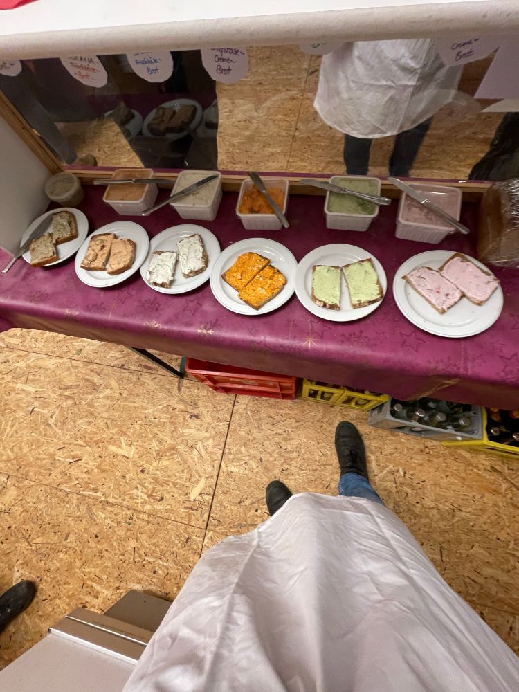 Man sieht meine Füße und eine Schürze (weiß) vor einem Stand mit mehreren Tellern auf denen Brote mit Aufstrich drauf sind 