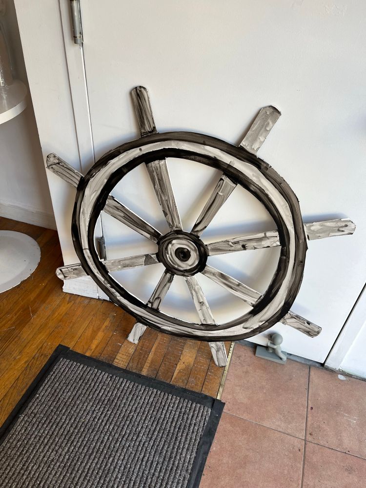 A ship’s wheel leans against the wall of a gallery waiting to be sited. It is white, outlined in black paint. By artist Helen Oliver Adelson. In front of it is a very ordinary black and white striped doormat. 
