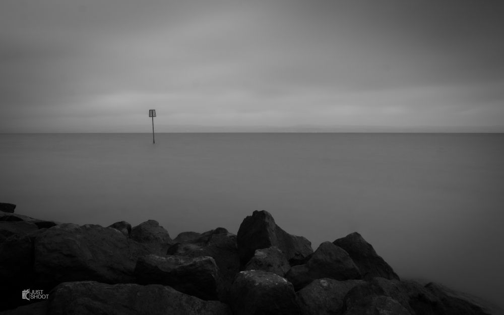 A black and white seascape photograph with rocks in the foreground, a singular metal object rising from the smooth water.