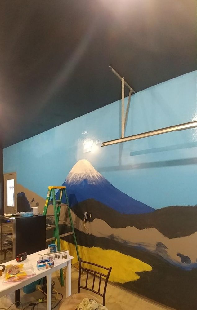 In progress photo of the Mt. Fuji mural. Mt Fuji has been painted with a Deep Blue base and a peak capped in snow that trails down the mountain and gradually fades. The sky in the background is a light Baby blue with no clouds yet. In the foreground, the shape of the forest of Coniferous Trees at the base of the mountain is nearly complete. The bright yellow ground of the valley has been painted, but the dirt has not been filled in yet, nor have any of the cherry blossom trees that the dirt surrounds.
●
Part of the left side of the mural is blocked by a green ladder and a black mini fridge with painting supplies on top. Towards the center of the picture there is a white, tall table with some cigarettes, paint cans, pretzels, and gloves scattered on top. There is a black and brown stool close to the perspective of the viewer. 