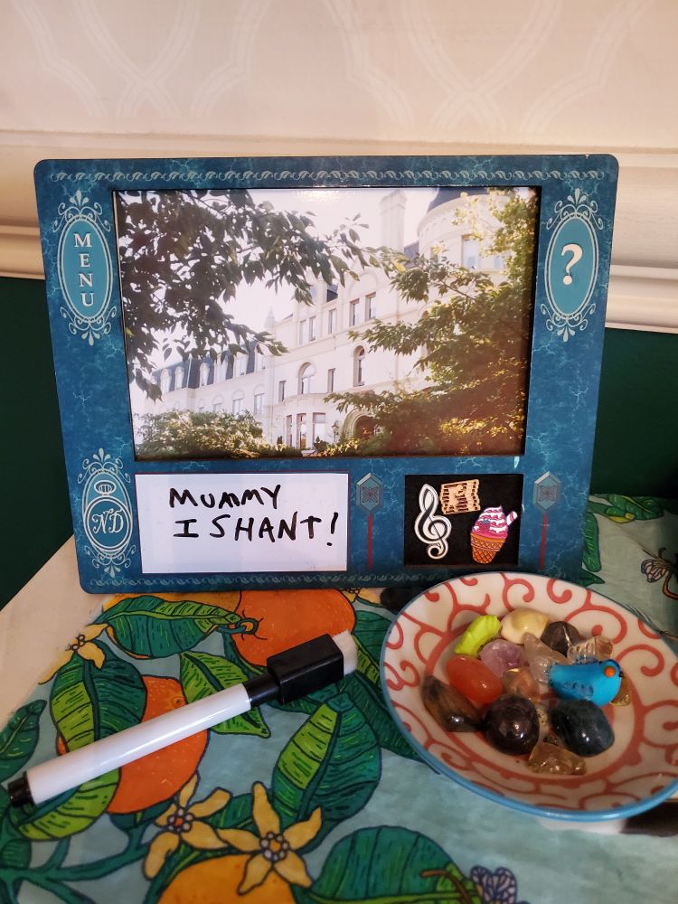 Image of a Blue/Teal picture frame that replicates the UI of early Nancy Drew games. The photo in the top center of the frame shows a multi-story building surrounded by trees. The left edge of the frame shows the Menu button and pocket watch with the initials ND, while the right edge includes the question mark and inventory slider buttons. 

The bottom half of the frame includes a space with a rectangular whiteboard for writing notes, and to the right of that there is a square section of black felt for displaying pins, which resembles the inventory of the old Nancy Drew games. Attached to the felt are 3 decorative pins: A treble clef, a strawberry ice cream cone, and a Nancy Drew pin that resembles the menu/logo of the earlier games: The pin is a stack of red/gold books with Nancy Drew's golden silhouette superimposed onto the left side of the stack. The whiteboard section currently reads in black marker, "Mummy I shan't!" 

The frame itself sits on a white mantle, which has a light blue tablecloth ran across it, with a design of oranges, blossoms, leaves and honeybees throughout. There is a Black marker/eraser sat on the mantle next to the frame, as well as a small red/white dish with a swirly design. The dish holds plenty of Earth candy (crystals, semiprecious stones) as well as a small glass statue of a Bluebird. 