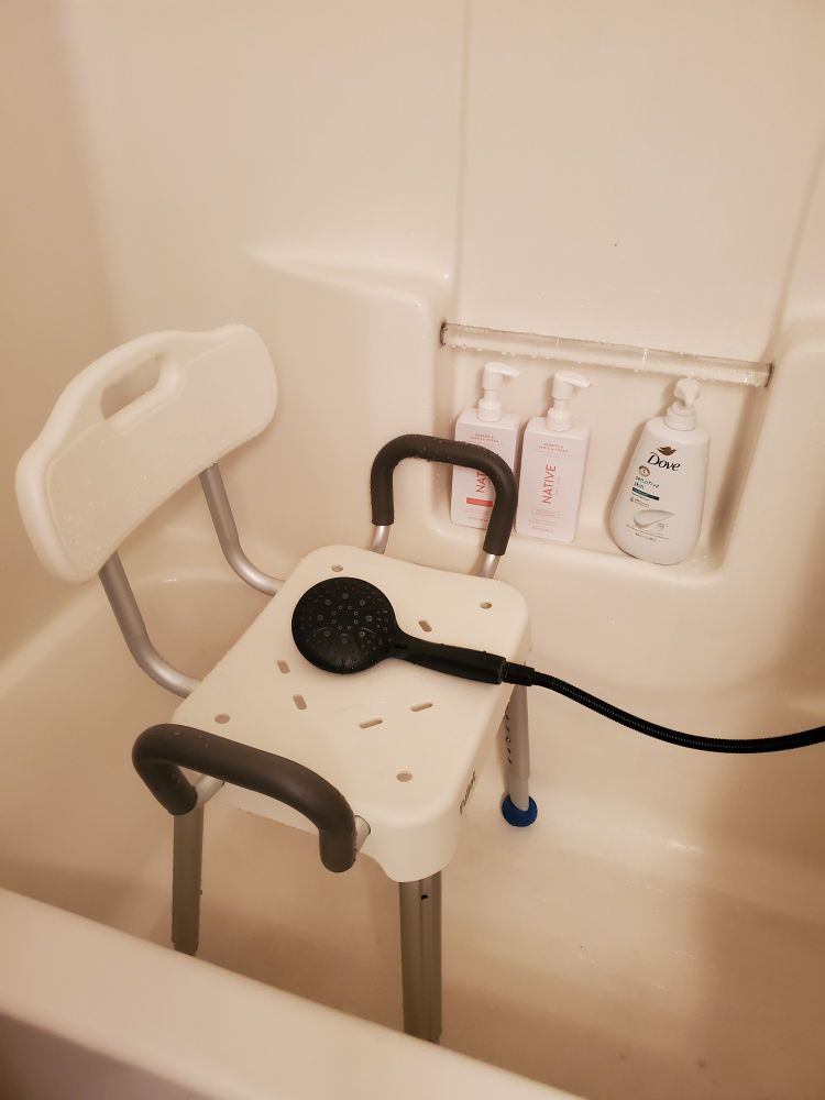 Image of a white shower/bath stall, with a white shower chair placed in the center of the tub near the back. The shower chair has a back and arms, and the arms have a grey grippy material on them for support. The chair also has multiple holes on the seat for water to drain.  There's a black detachable shower head placed on the seat, with the hose attached out of frame. The stall has a cubby for placing items in the center with a rod to hang a rag on. There's soap by DOVE and Native placed in the cubby.