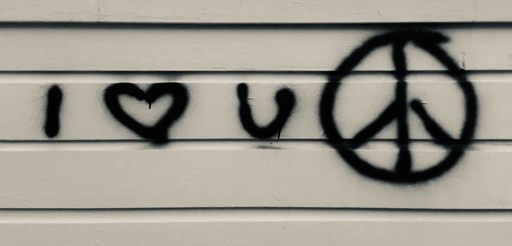 Auf einem weißen Bretterzaun steht in schwarzer Lackfarbe: I Herz u Peacezeichen (I love you peace).