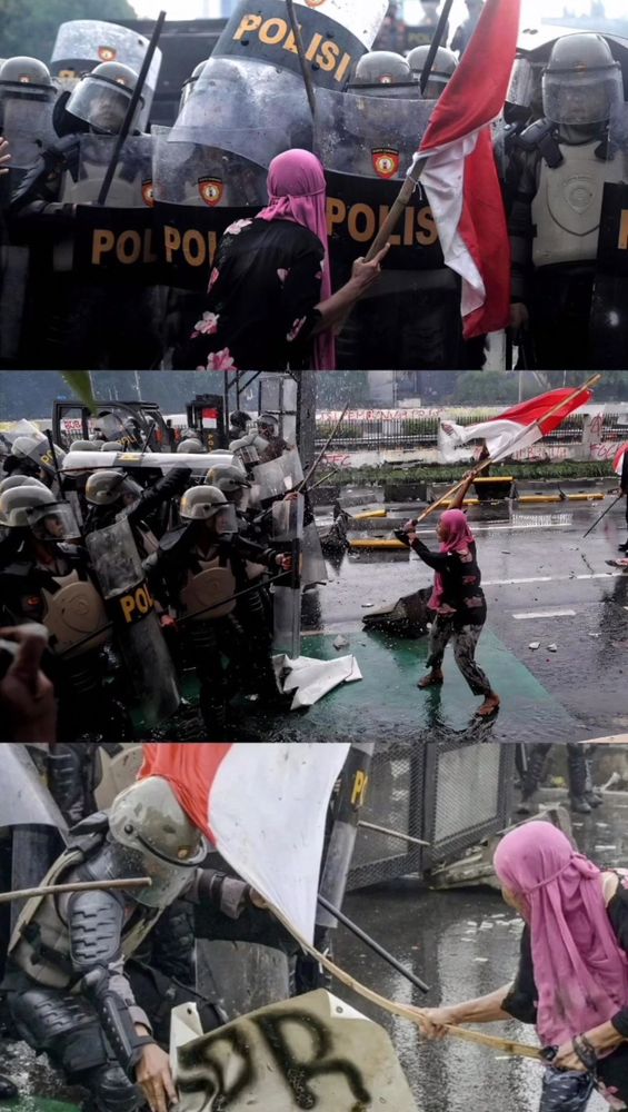 A woman in a pink hijab and holding the Indonesian flag attached to a pole is leading the front of rioters against a police shield barricade