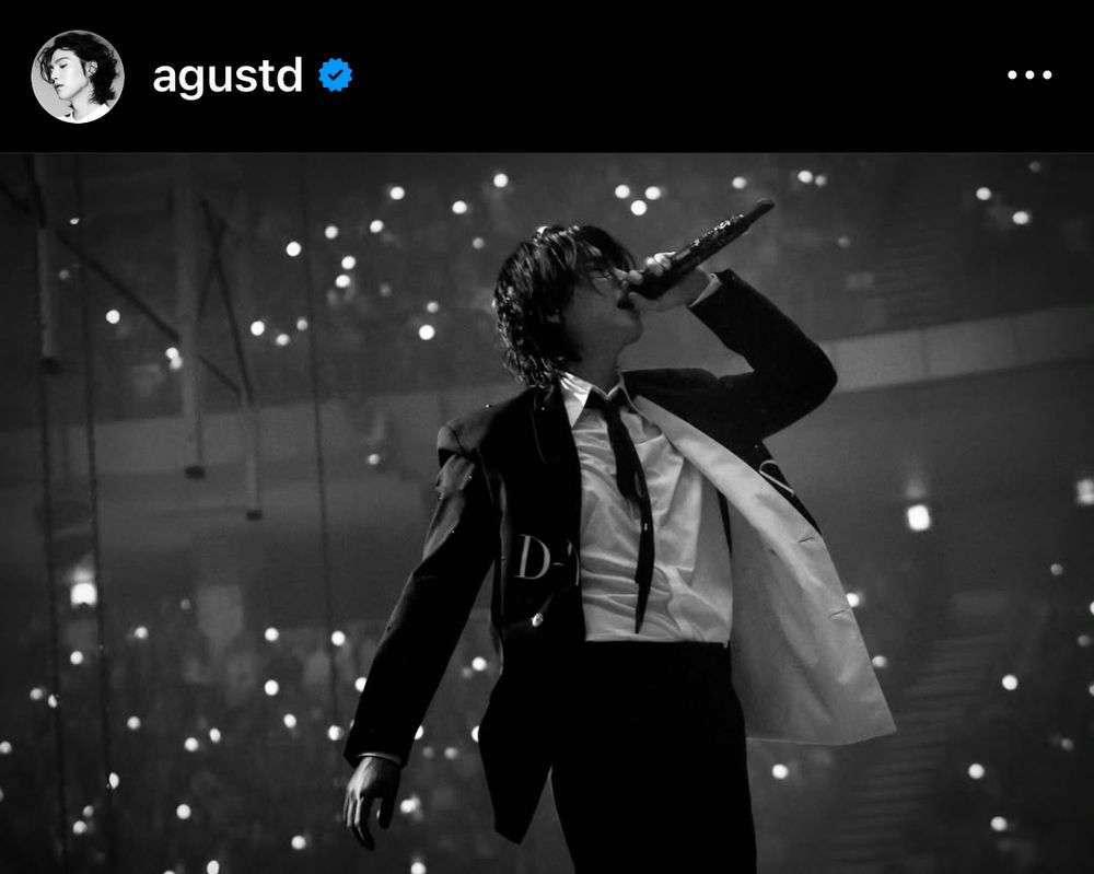A black and white photo of an Asian man on stage [SUGA of BTS], wearing  a suit and tie, white shirt. He was holding his mic with his left hand.
On the background, fans at the concert.
