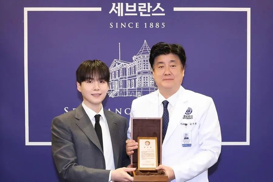 Two Asian men [Left: SUGA of BTS and Director Geum Ki-chang of Yonsei Medical Center] holding a plaque in front of Severance Hospital purple banner