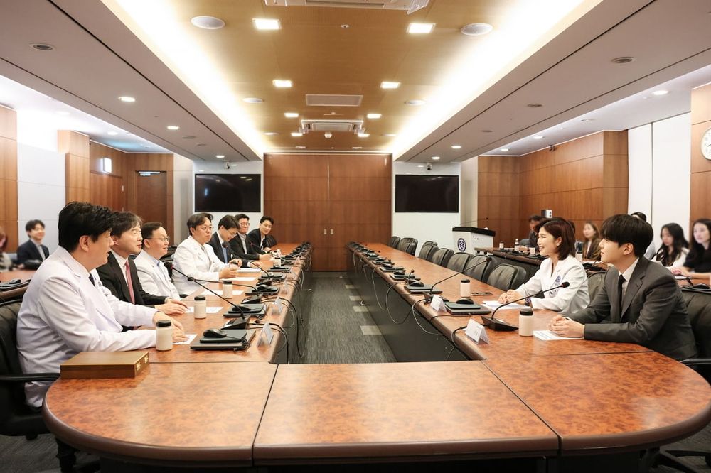 Wide shot of a meeting room with wooden desks. 
SUGA of BTS sat on the right side wearing medium brown suit