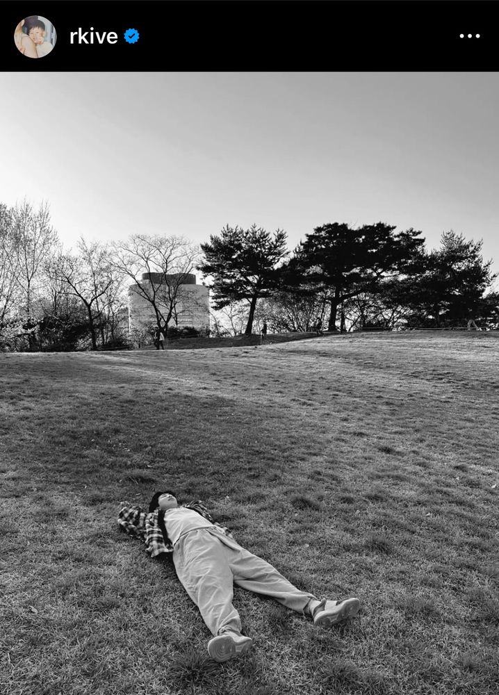 A black & white photo of an Asian man [RM of BTS] laying down on a grassy field.