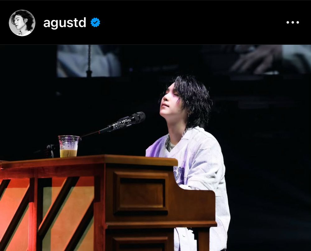 An Asian man [SUGA of BTS] playing a wooden piano on stage. He was closing his eyes