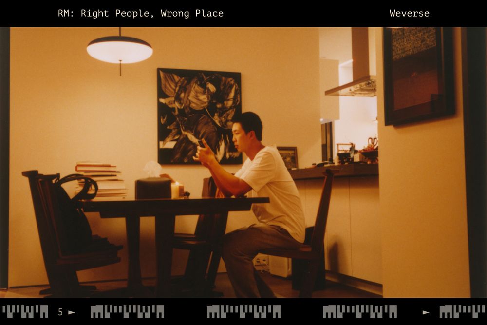 A sepia photo of an Asian man [RM of BTS] sitting at a dining area. On the background, a painting