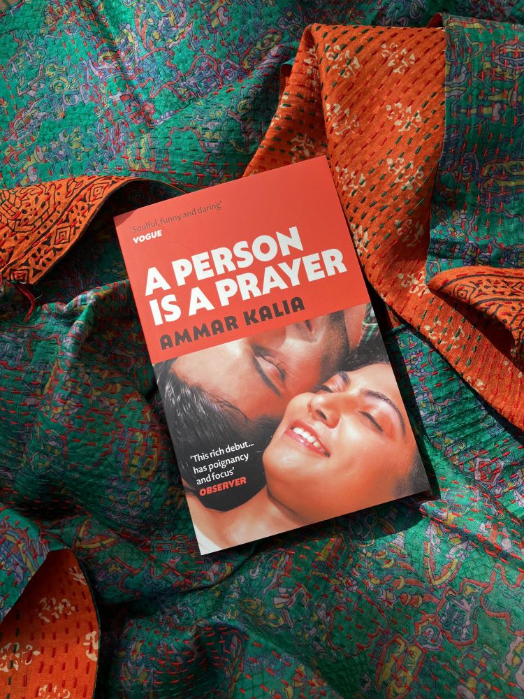 The book 'A Person Is A Prayer', with the title in bold white text in the upper half, the author's name Ammar Kalia beneath that, then a photo of two brown skinned people smiling with their eyes closed in the lower half of the cover. It sits on a multi-coloured fabric background of mixed turquoise and orange. 
