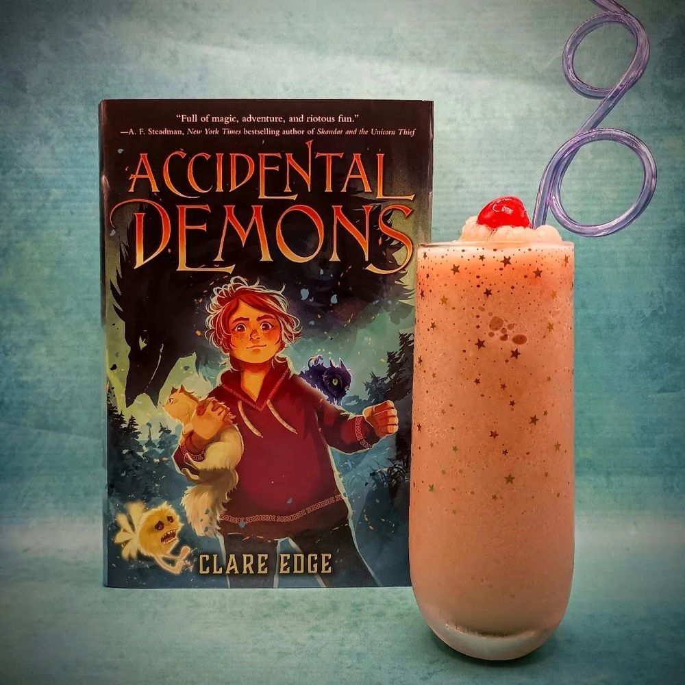 A photo of the physical book of Accidental Demons by Clare Edge, next to an orange smoothie I'm a glass with gold stars, topped with a cherry and with a purple silly straw
