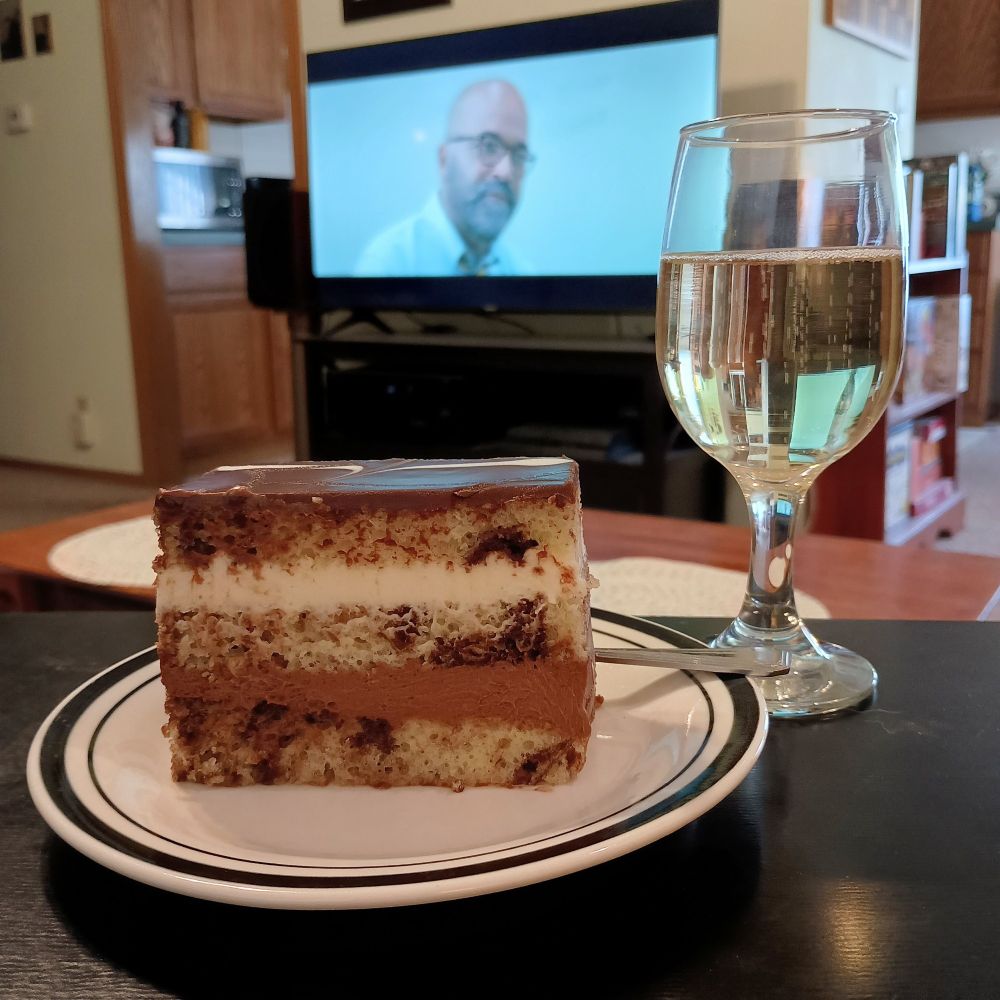 A photo of a layered chocolate and vanilla cake on a plate, next to a glass of sparkling wine. 

In the background a TV shows the opening frames of the movie American Fiction - the actor Jeffrey Wright standing against a light blue background.