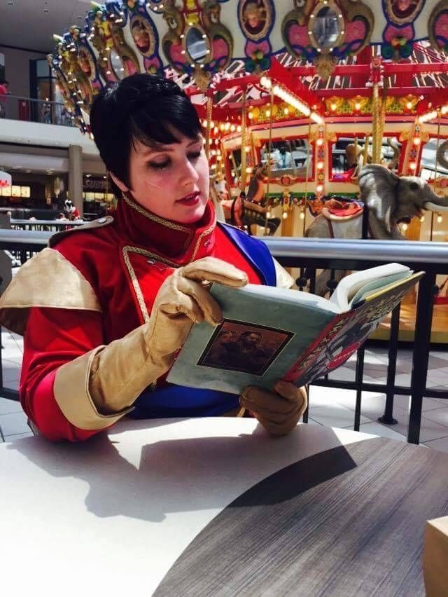 Amy as Cassandra from Dragon Age 2 and Inquisition. She reads a copy of Varric's book. Short dark hair and red formal jacket.