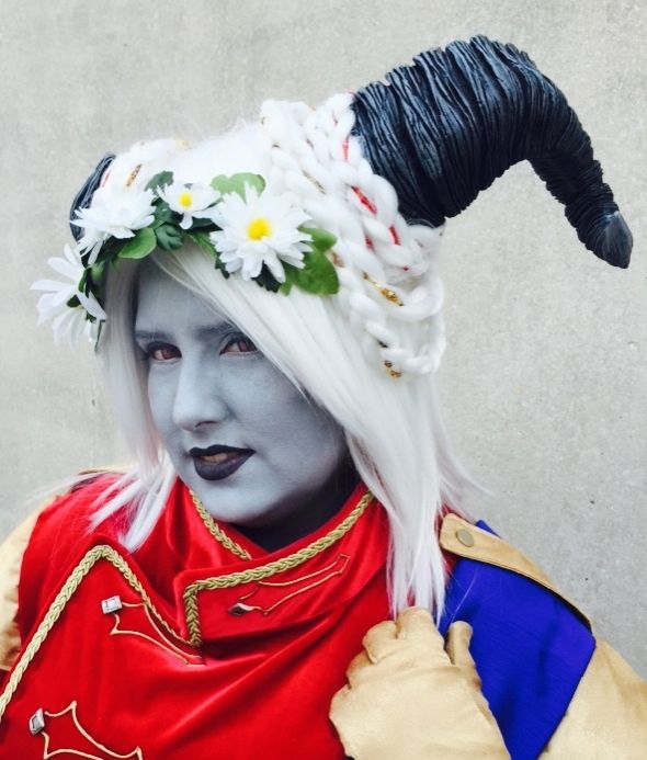 Amy as a Qunari Inquisitor from Dragon Age, in cosplay. Gray skin, black horns, and white wig with formal red jacket and a white daisy flower crown.