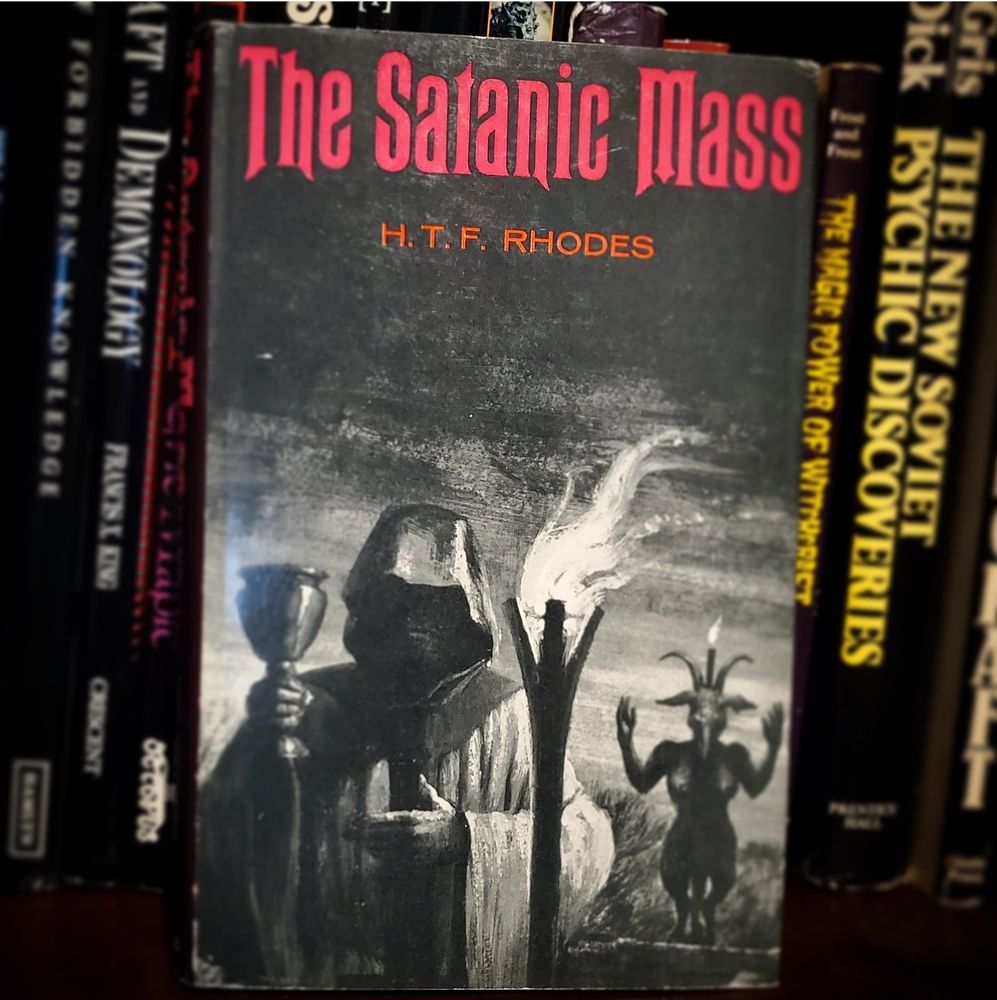 A book cover featuring a robed and hooded figure holding a chalice next to a torch and statue of a humanoid goat, titled The Satanic Mass, by H. T. F. Rhodes.