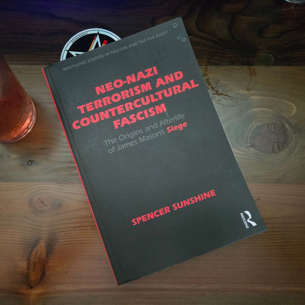 A book on a wooden surface, titled Neo-Nazi Terrorism and Countercultural Fascism: The Origins and Afterlife of James Mason's Siege, by Spencer Sunshine