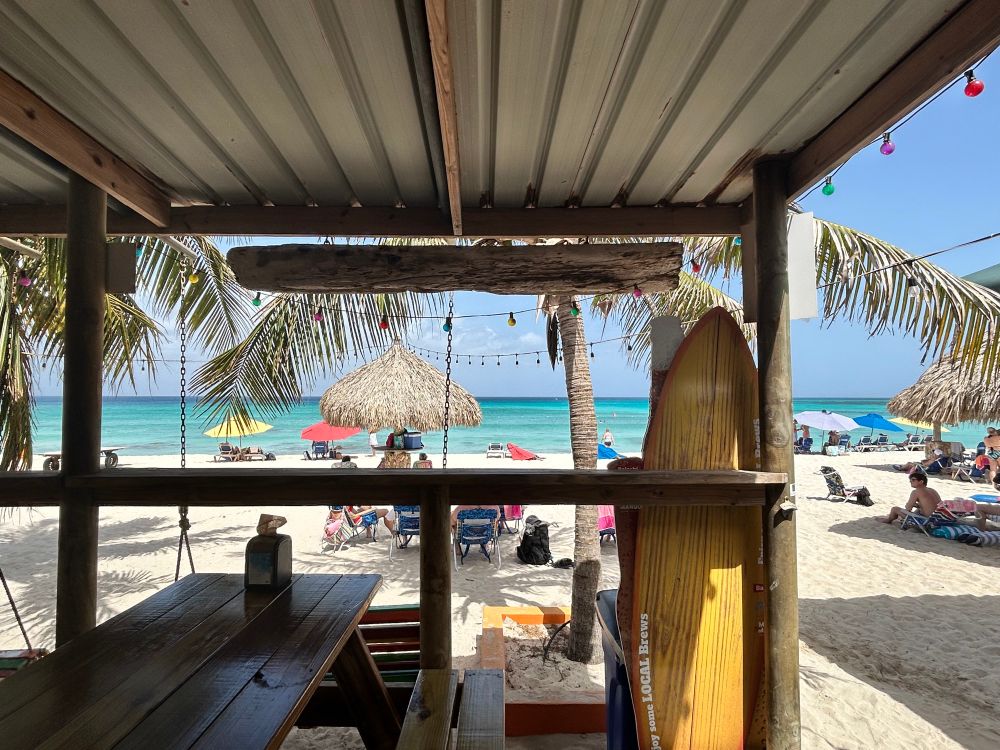 Looking out from inside a beach shack restaurant. Surfboard to the right, sand, palapa a, and the aquamarine colors of the Caribbean. 