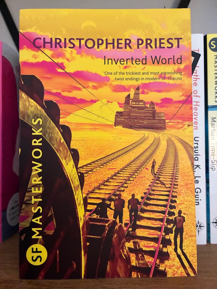 Cover image of the SF Masterworks edition of ‘Inverted World’ by Christopher Priest, depicting a large pulley system with cables reaching out across a series of rail tracks - in the distance, the cables are attached to a ‘mobile’ city. A group of workers stand around the tracks monitoring the movement of the city as it is pulled along the track system.