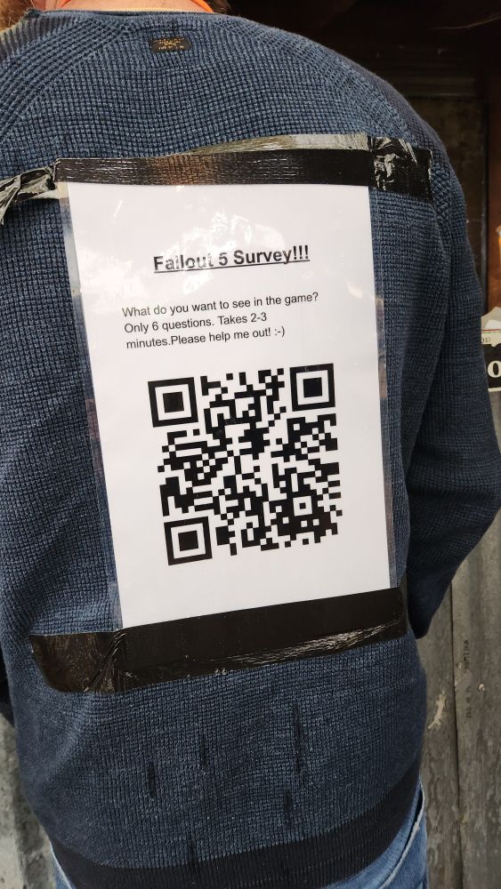 Man with a press badge has a sign duct taped to his back. A QR code is on the sign with text that reads: "Fallout 5 Survey!!! What do you want to see in the game? Only 6 questions. Takes 2-3 minutes. Please help me out! :-)"
